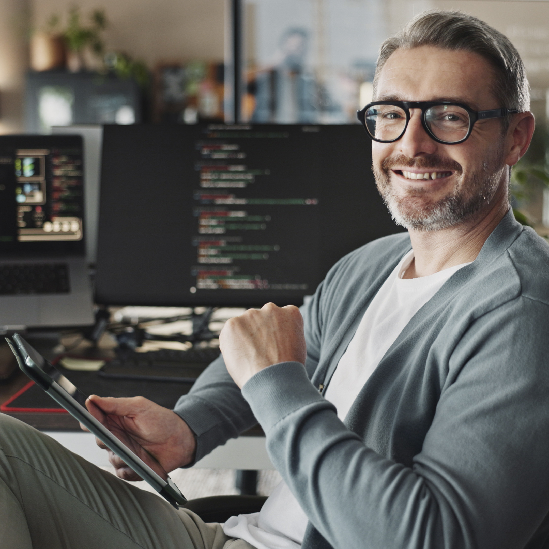 Ein Mann arbeitet an Computercodes an zwei Bildschirmen und nutzt dabei ein Tablet, während er lächelt.