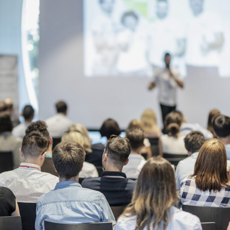 Ein Mann steht vor einem Publikum und hält einen Vortrag, im Hintergrund eine Leinwand.
