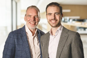 Ein Bild von Alexaner und Nicolas Verweyen in Business-Outfits, die zusammen in einem modernen Büro lächeln.